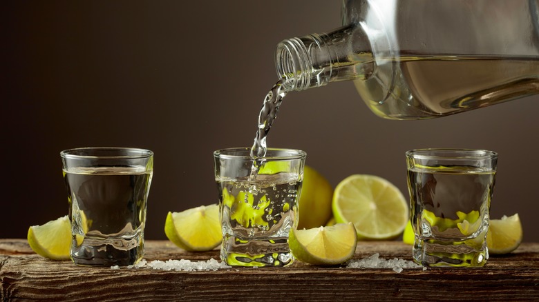 A bottle filling shot glasses with tequila on a board with sliced limes