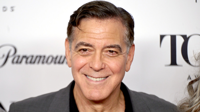 Actor George Clooney smiling and wearing a gray suit jacket with a black shirt