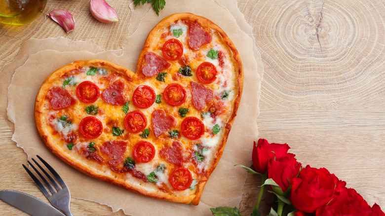 A heart-shaped pepperoni pizza on a brown table, with red onions and red roses